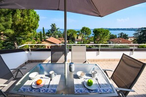 A glass table with a view of a coastal landscape, two chairs, and a table setting with plates, a bowl, a spoon, a fork, and a cup. - Cupido (Gardone Riviera)