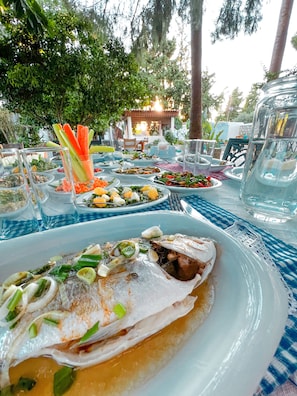Una mesa de comedor con diversos platos, incluyendo pescado y verduras, dispuesta al aire libre con árboles al fondo.