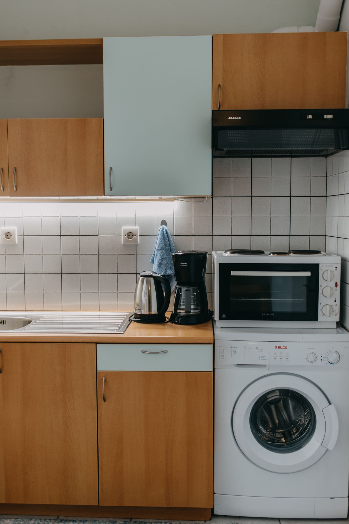 A compact kitchen with wooden cabinets, a built-in oven, a coffee maker, and a front-loading washing machine.