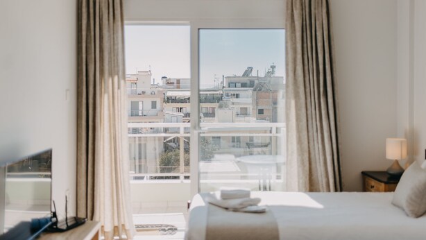 A hotel room with a bed, a desk, a chair, a window with curtains, and a view of buildings outside. - Best House Town View Apartment (Patras)