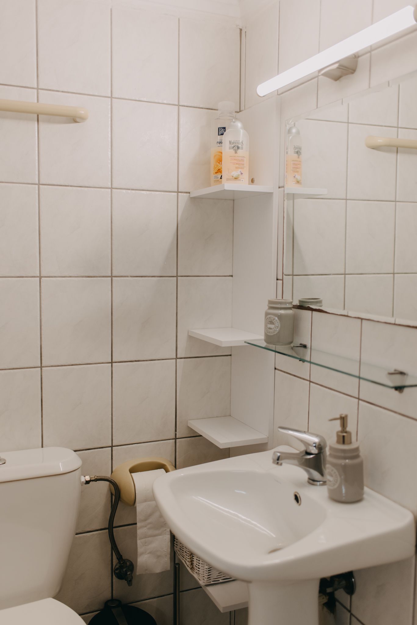 Un baño con azulejos blancos, un lavamanos blanco, un espejo y estantes con artículos de aseo.