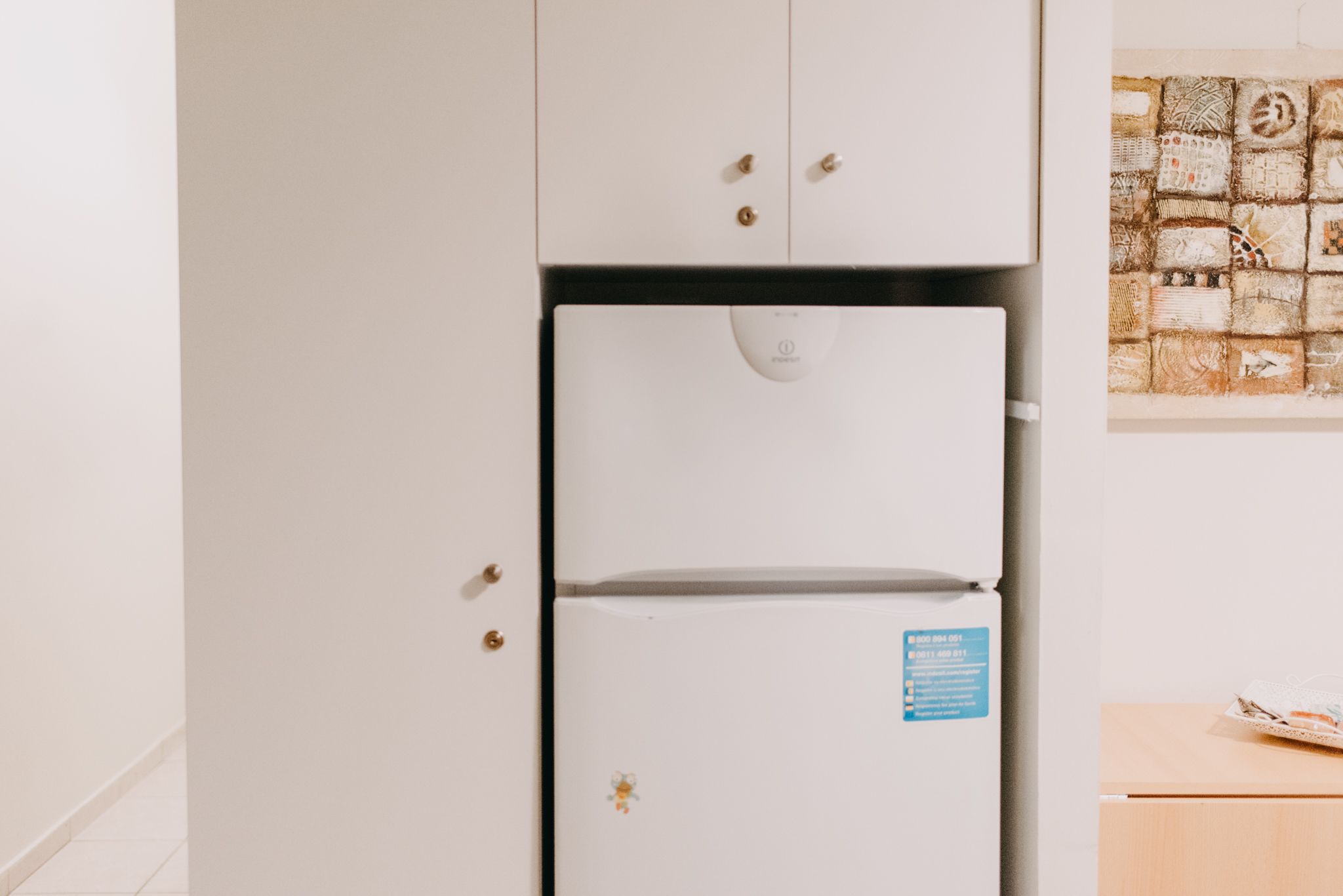 A white refrigerator with a blue sticker, a white cabinet above it, and a wall with a textured art piece.