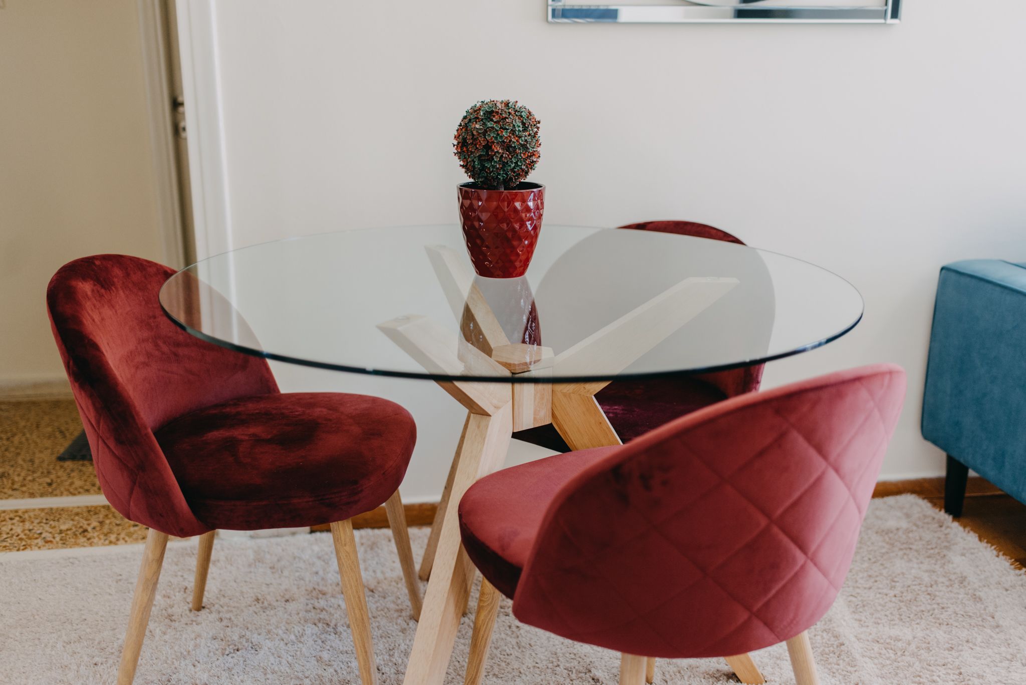 A round glass table with a red potted plant, surrounded by two red velvet chairs with wooden legs.