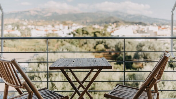 Un balcón con una mesa de madera y dos sillas, con vistas a un pueblo y montañas al fondo.