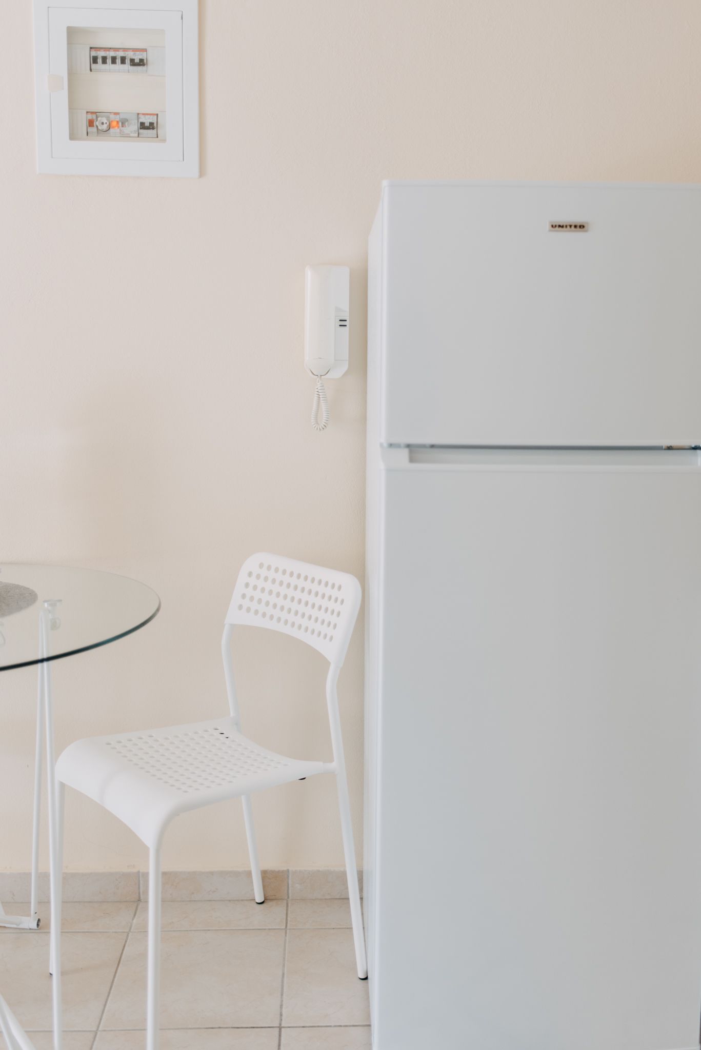 A compact kitchen with a white refrigerator, a small round glass table, and a white plastic chair.