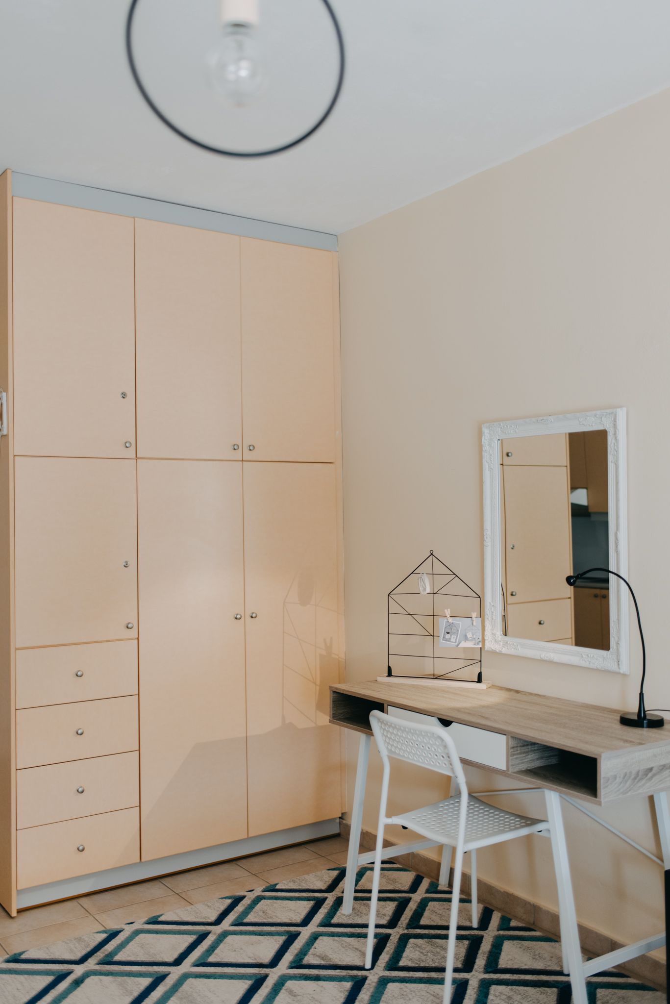 A room with a large wardrobe, a desk with a chair, a mirror, and a geometric-patterned rug.
