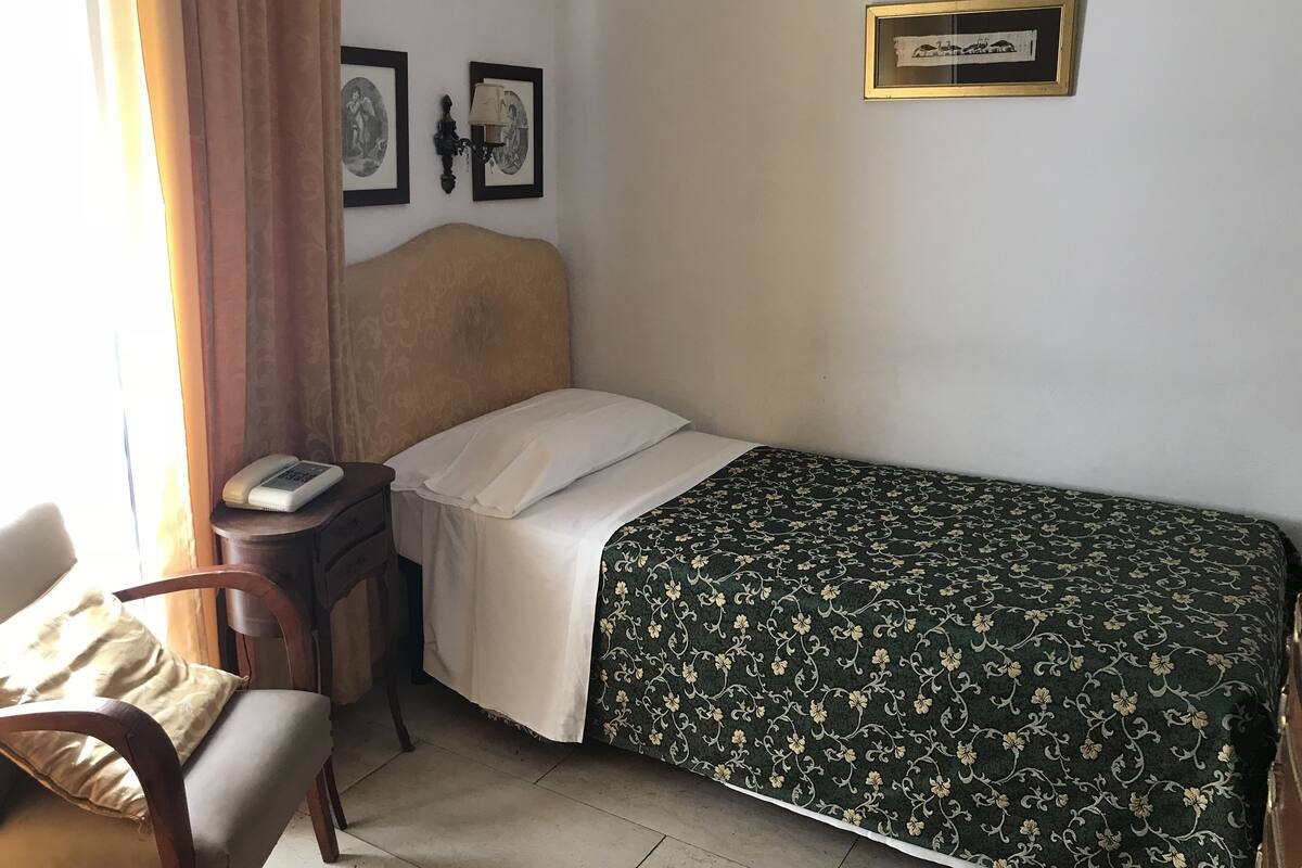 Un dormitorio pequeño y ordenado con una cama individual, una mesita de noche con lámpara, una silla y dos cuadros en la pared.