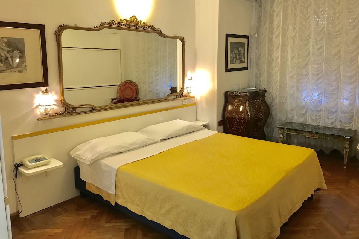 Habitación de hotel con cama, mesita de noche con teléfono, espejo y un jarrón decorativo.