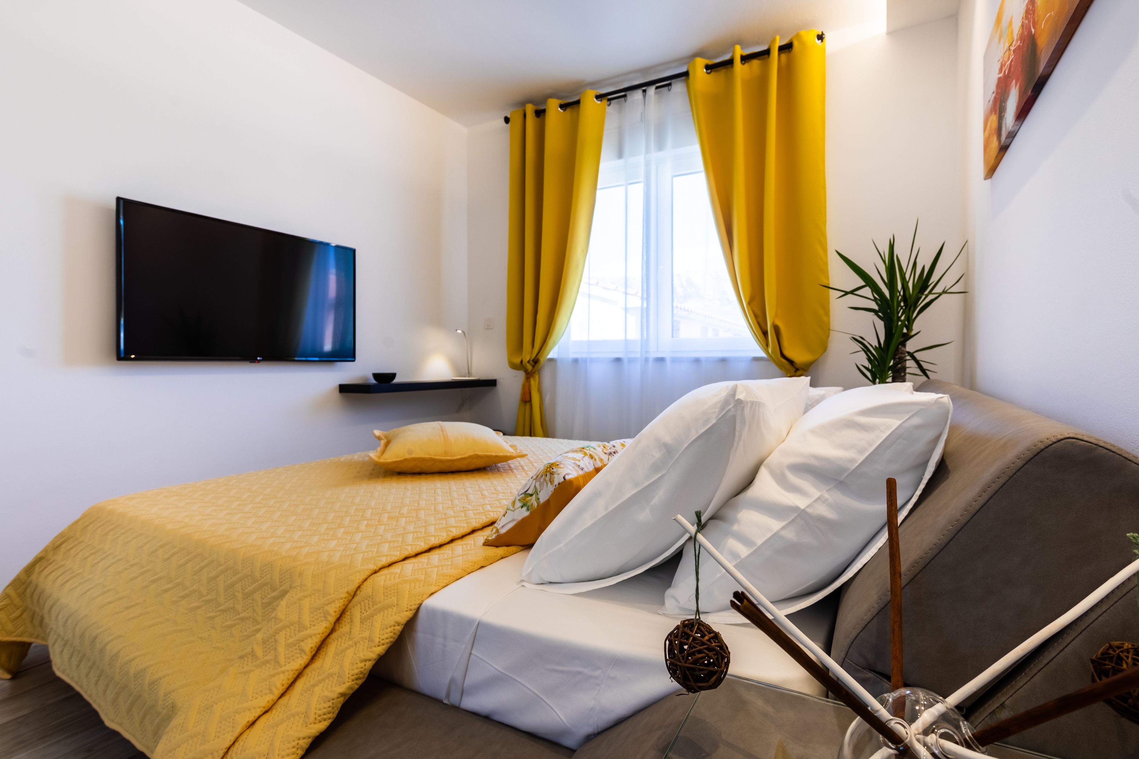 A modern bedroom with a bed, a flat-screen TV, a window with yellow curtains, and a plant.