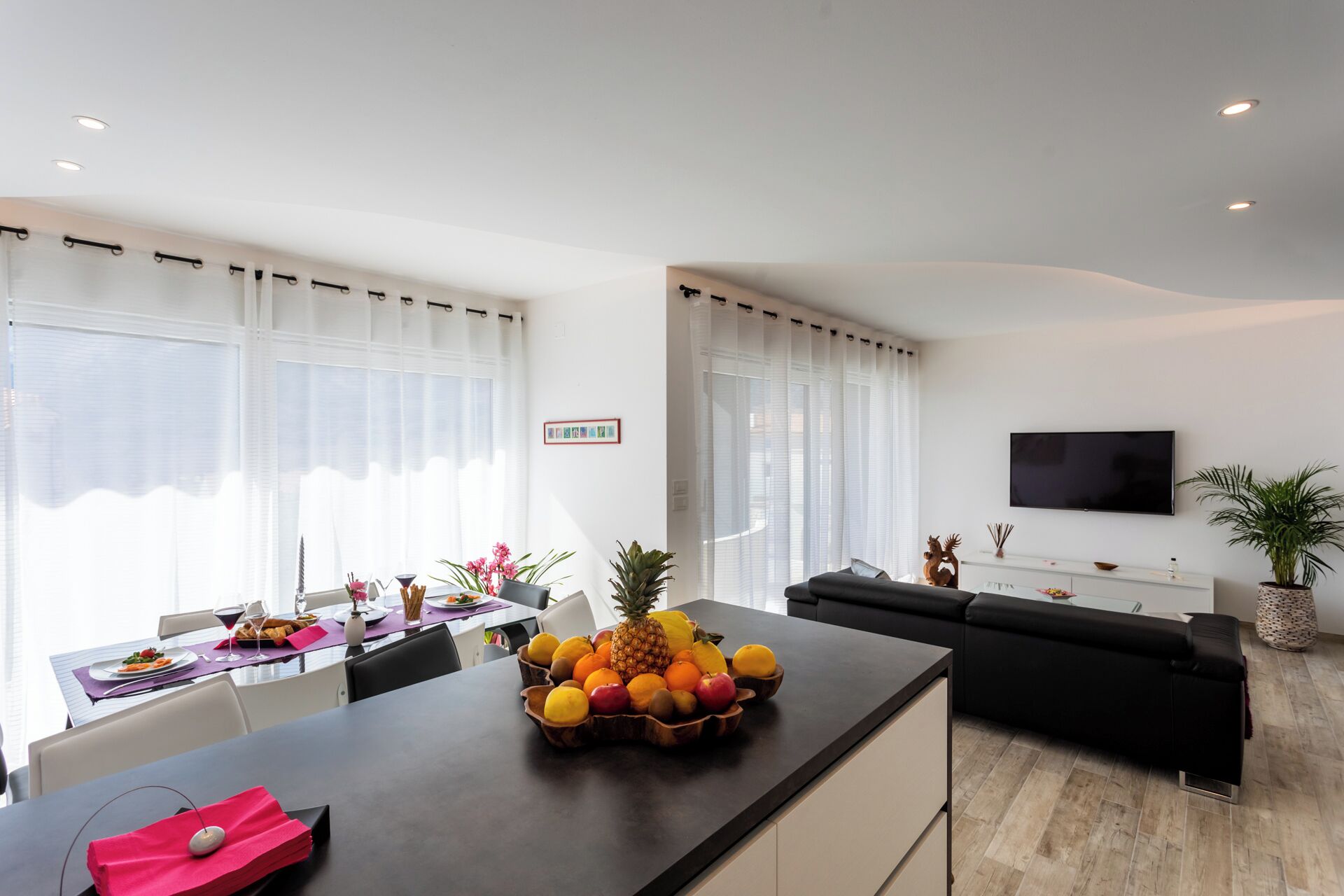 Una sala moderna con un sofá negro, un comedor con una mesa preparada para dos personas, y un frutero en la encimera de la cocina.