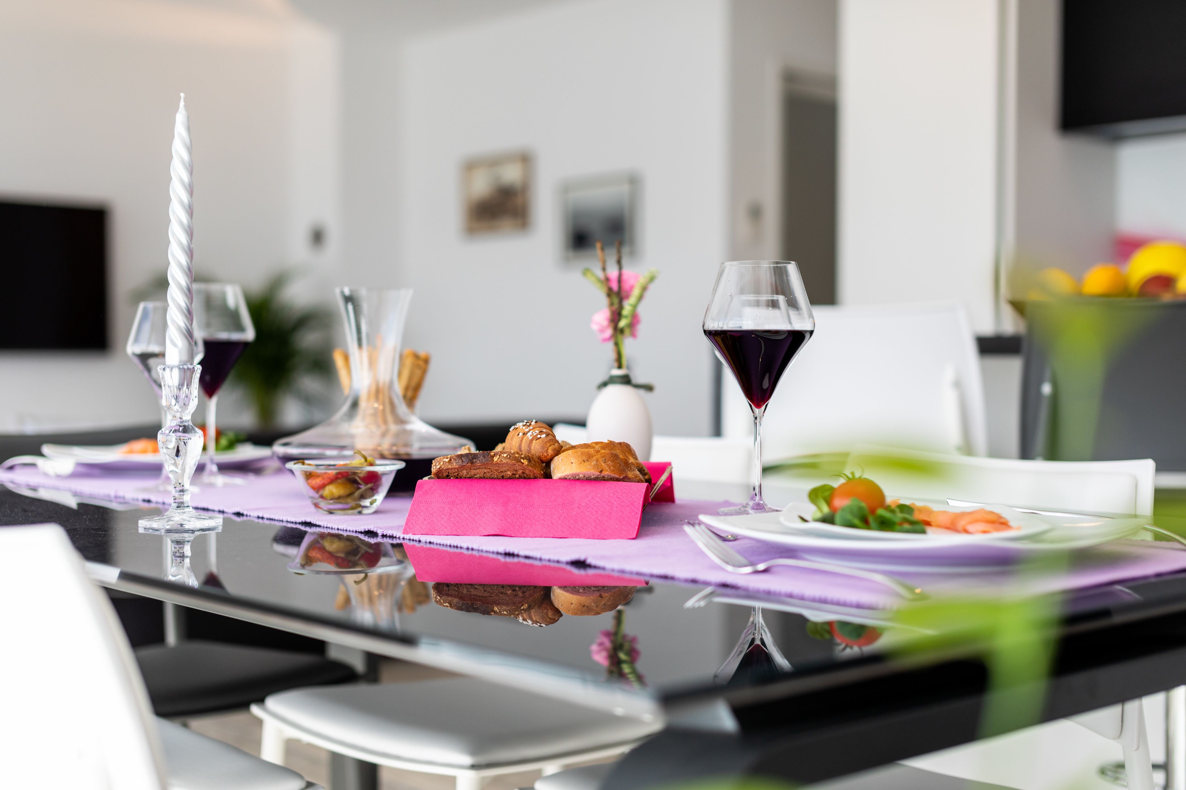 A dining table with a pink placemat, a glass candle, a glass of red wine, a bowl of pastries, and a vase with flowers.