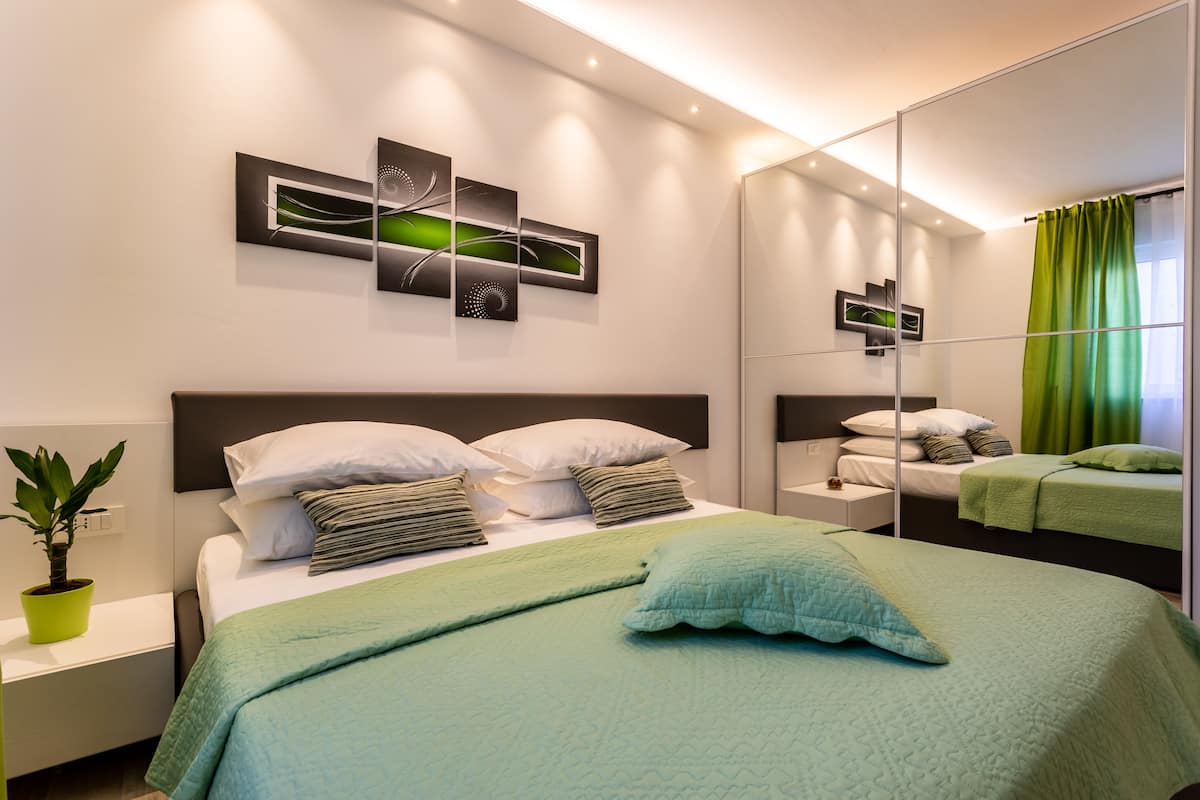 A modern bedroom with a large bed, a bedside table with a plant, and a mirror reflecting the room.