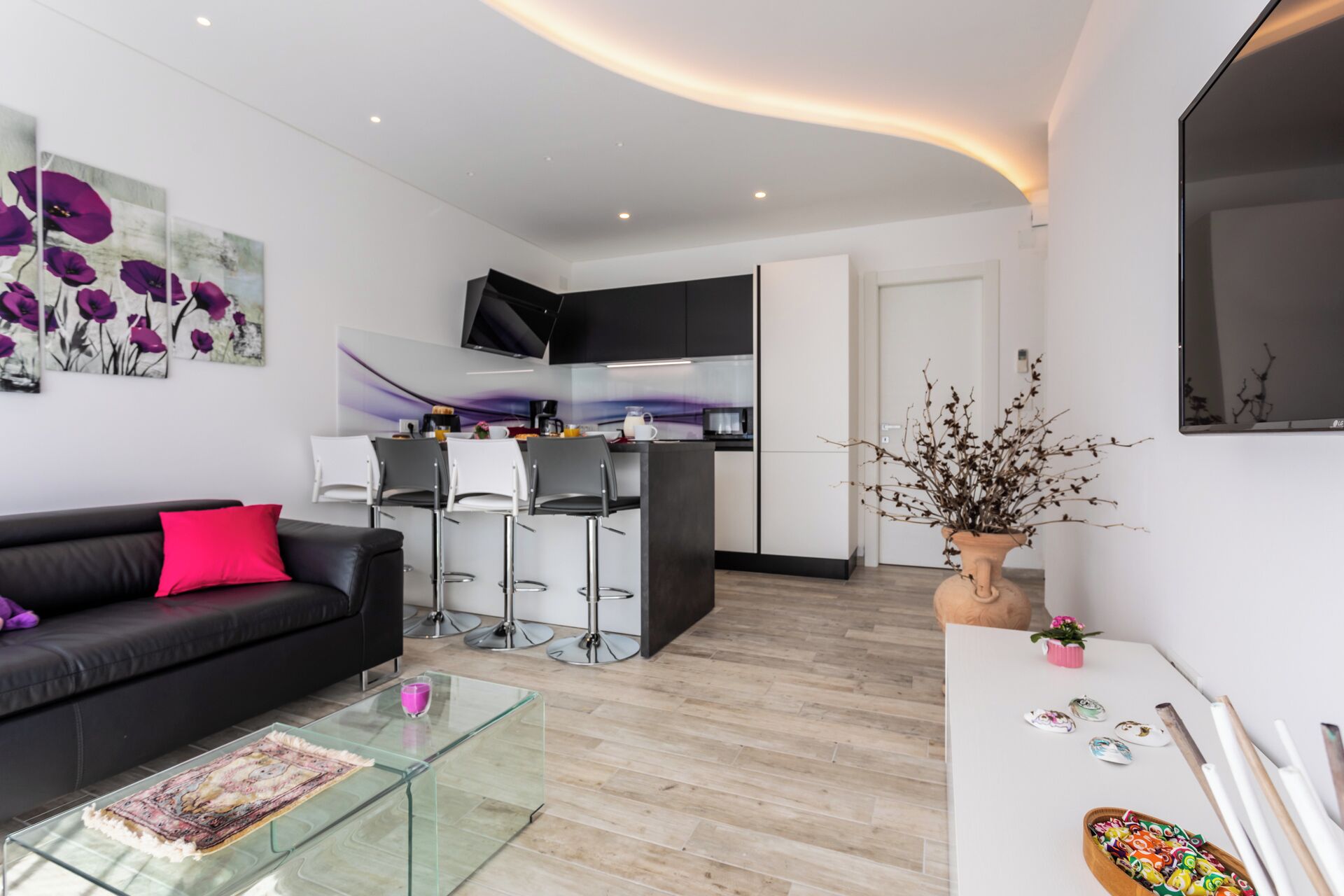A modern living room with a black sofa, a glass coffee table, and a kitchen area with a TV above the counter.