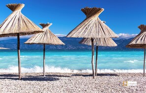 A beach with pebbled shore, clear turquoise water, and thatched umbrellas. - Villa Valis (Baska)