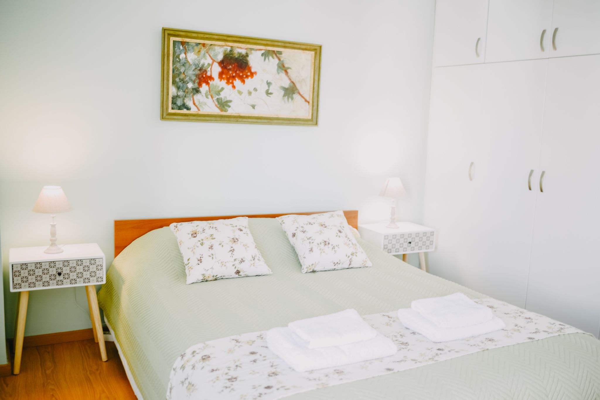 A neatly arranged bedroom with a bed, two bedside tables, a nightstand, a wardrobe, and a framed picture on the wall.