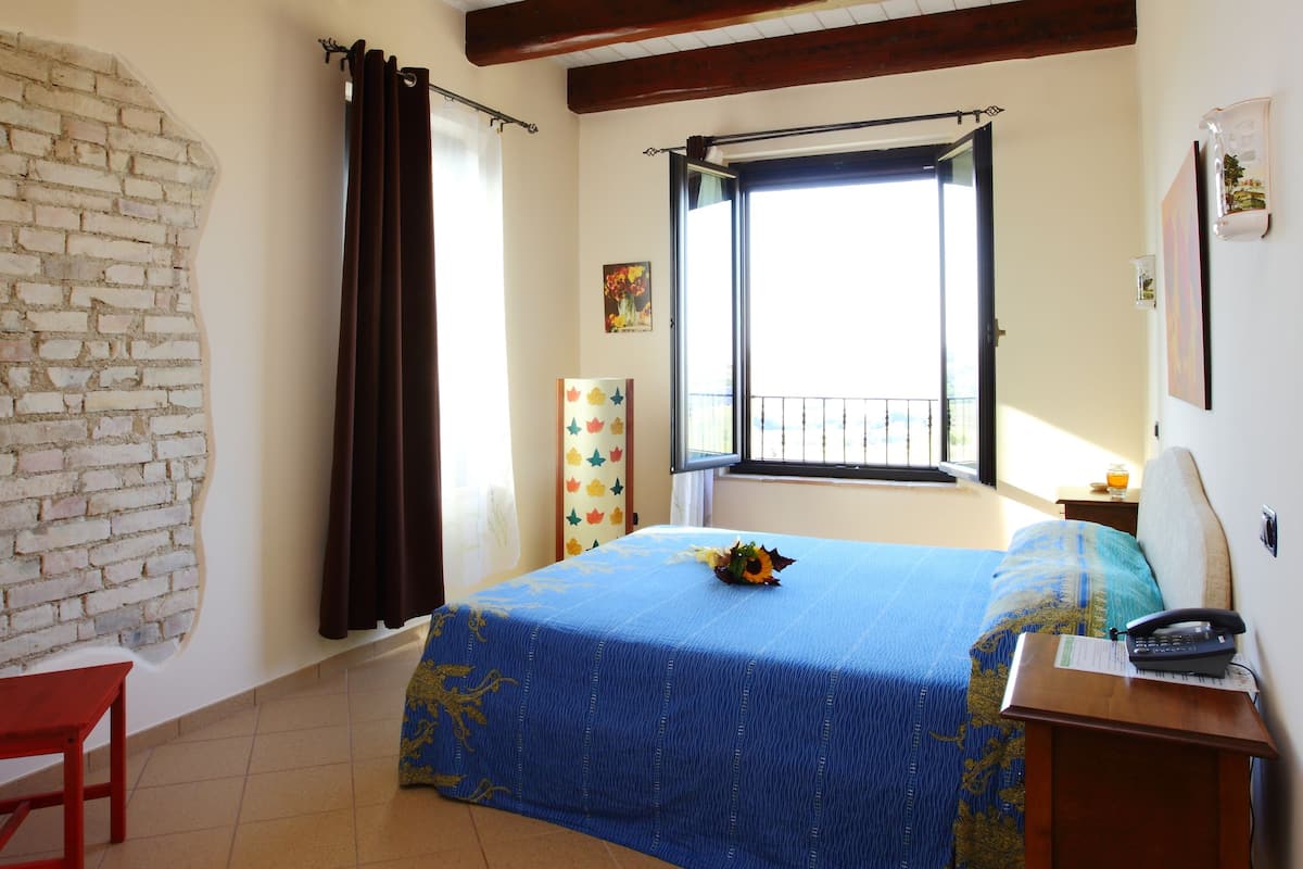 Un dormitorio con una cama, una mesa de madera, una silla, un ventanal con cortinas y un balcón.