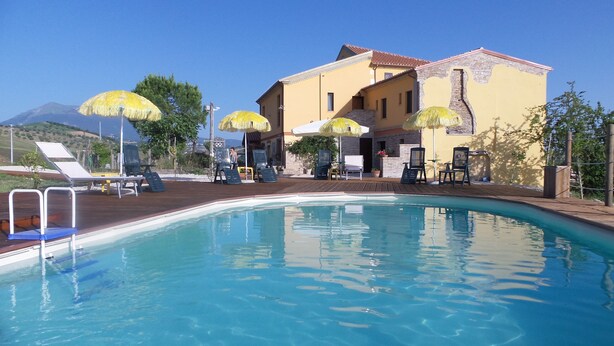 A pool area with lounge chairs and umbrellas, a building with a tiled roof, and a mountain in the background.