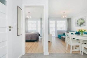 Um apartamento moderno com quarto visível, área de jantar com mesa e cadeiras brancas, e sala de estar com sofás azuis-esverdeados.
