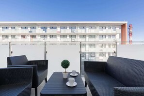 Une terrasse sur le toit, meublée de mobilier en osier, avec une petite table, des tasses et une plante en pot, et, en arrière-plan, un bâtiment blanc doté de balcons.