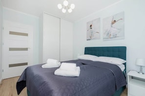 Un lit bien fait, recouvert d’une couvre-lit bleu foncé et agrémenté d’oreillers blancs, avec une tête de lit turquoise et deux tableaux représentant des danseurs, accrochés au mur.