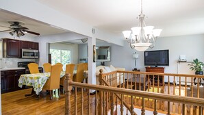 A spacious kitchen with wooden cabinets, a dining area with yellow chairs, a staircase with a wooden railing, and a living room with a TV and a plant.