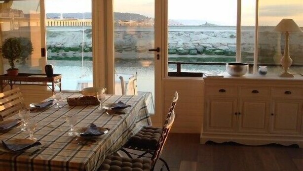 Un coin repas avec une table dressée pour deux personnes, un meuble blanc et une vue sur une côte rocheuse, visible à travers de grandes fenêtres.