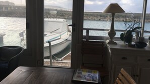 Une pièce avec vue sur un bateau amarré dans une marina, une table en bois avec un journal, une lampe et une plante en pot.