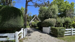 Une allée de gravier mène à une maison entourée d’une clôture en bois peinte en blanc, le tout étant bordé d’une végétation luxuriante et d’arbres.