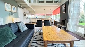 Un salon moderne avec un canapé noir, une table basse en bois et un tapis à motifs zébrés. On aperçoit, au fond, un coin cuisine avec un store rouge à la fenêtre.