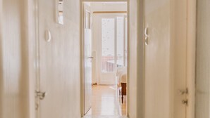 A narrow hallway with white walls and a speckled floor, leading to a room with a large window and a door. - Best House Attiki Metro (Athens)