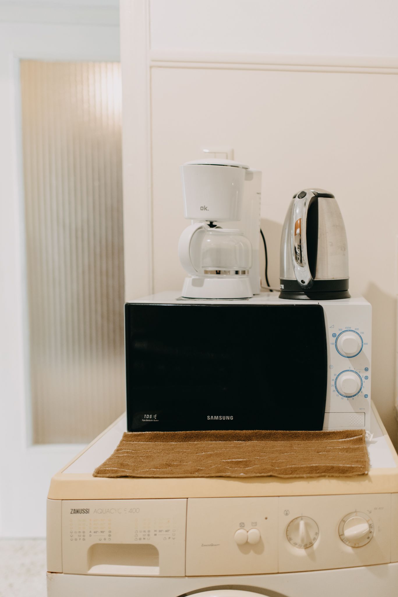 Un microondas Samsung sobre un electrodoméstico blanco, con una cafetera y un hervidor encima.