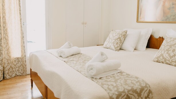 A neatly made bed with white linens and decorative pillows, a wooden headboard, and a window with curtains. - Best House Attiki Metro (Athens)