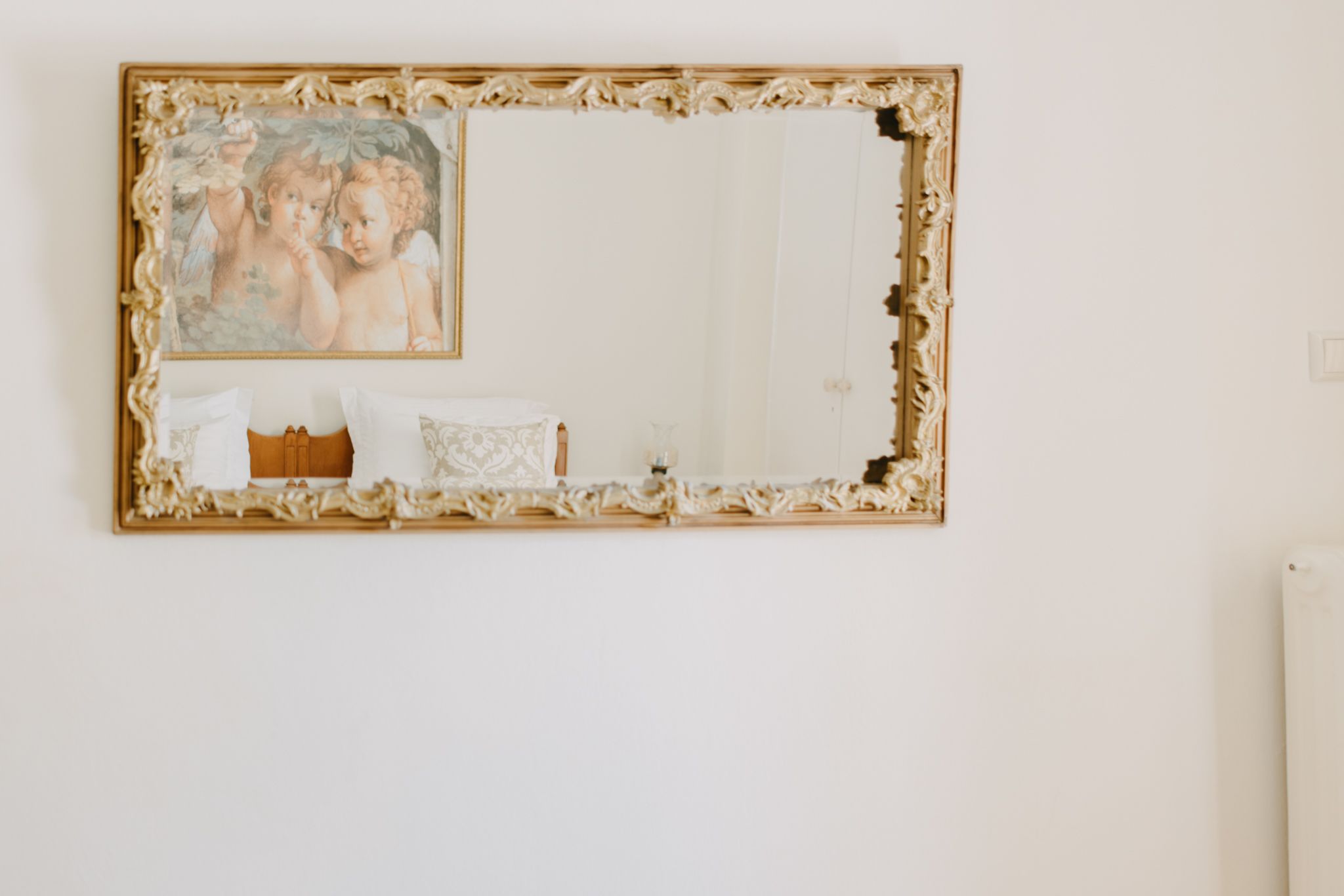 Un espejo grande y ornamentado con un marco floral, una mesita con mantel blanco y un cuadro de querubines en la pared.