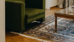 A room with a green armchair, a patterned rug, a wooden coffee table, a door with a white frame, and a window with blinds. - Best House Attiki Metro (Athens)