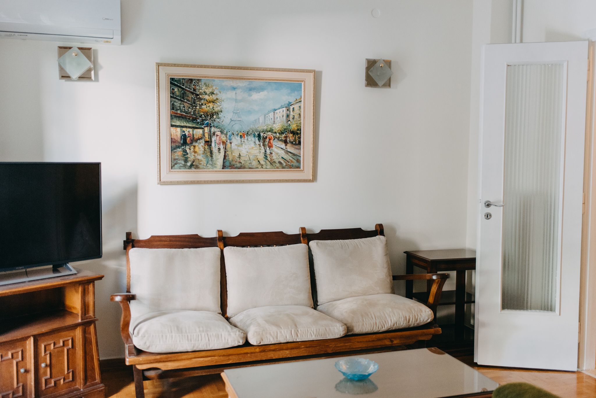 A living room with a wooden sofa, a television, a painting of a street scene, and a door.