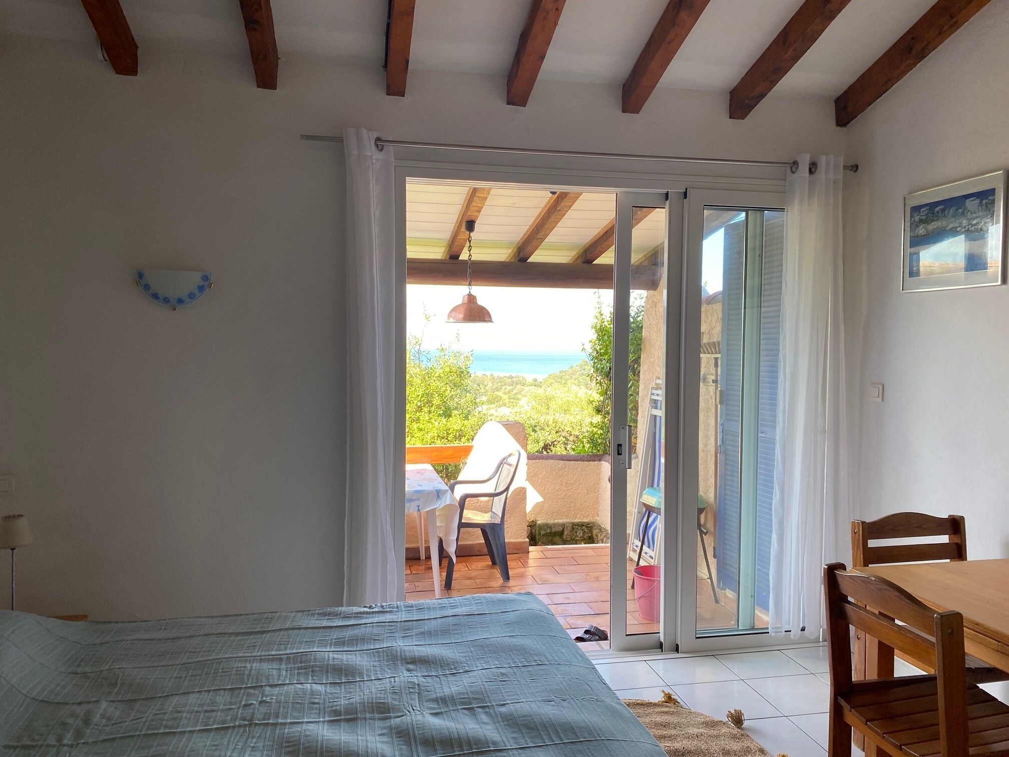 A bedroom with a bed, a wooden table, and a chair. There is a sliding glass door leading to an outdoor area with a view of the sea.