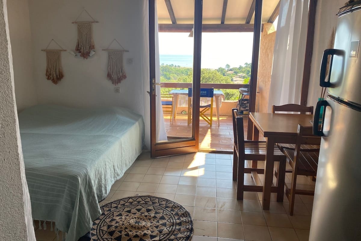 A room with a bed, a dining table with chairs, a refrigerator, and a view of the outdoors.