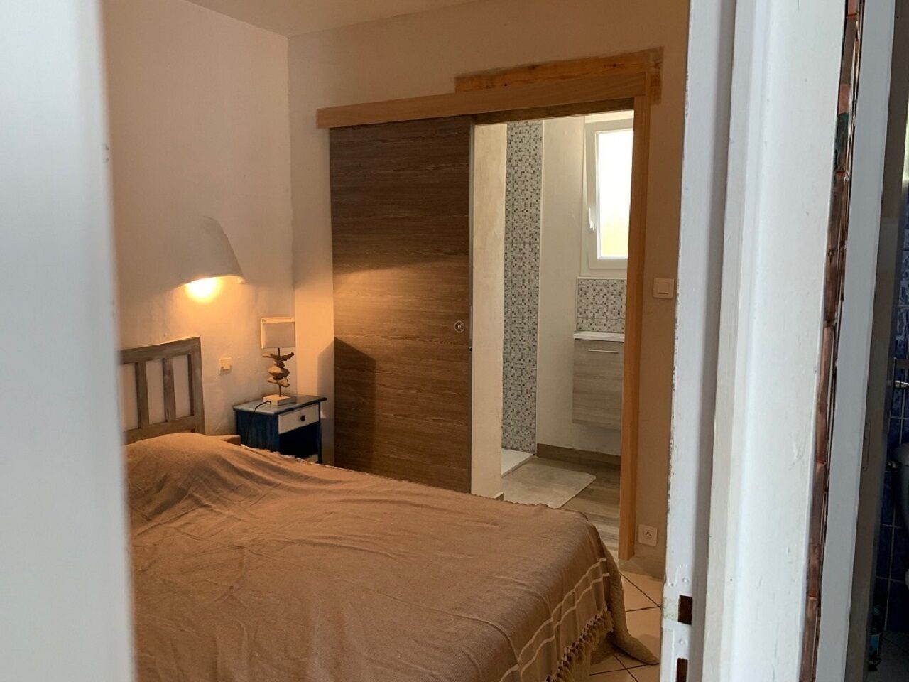 Een slaapkamer met een bed, nachtkastje, lamp en een houten deur naar een badkamer.