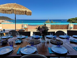 A table set for dining with a view of the sea, a thatched umbrella, and wicker chairs. - Residence Fium del Cavo (Zonza)