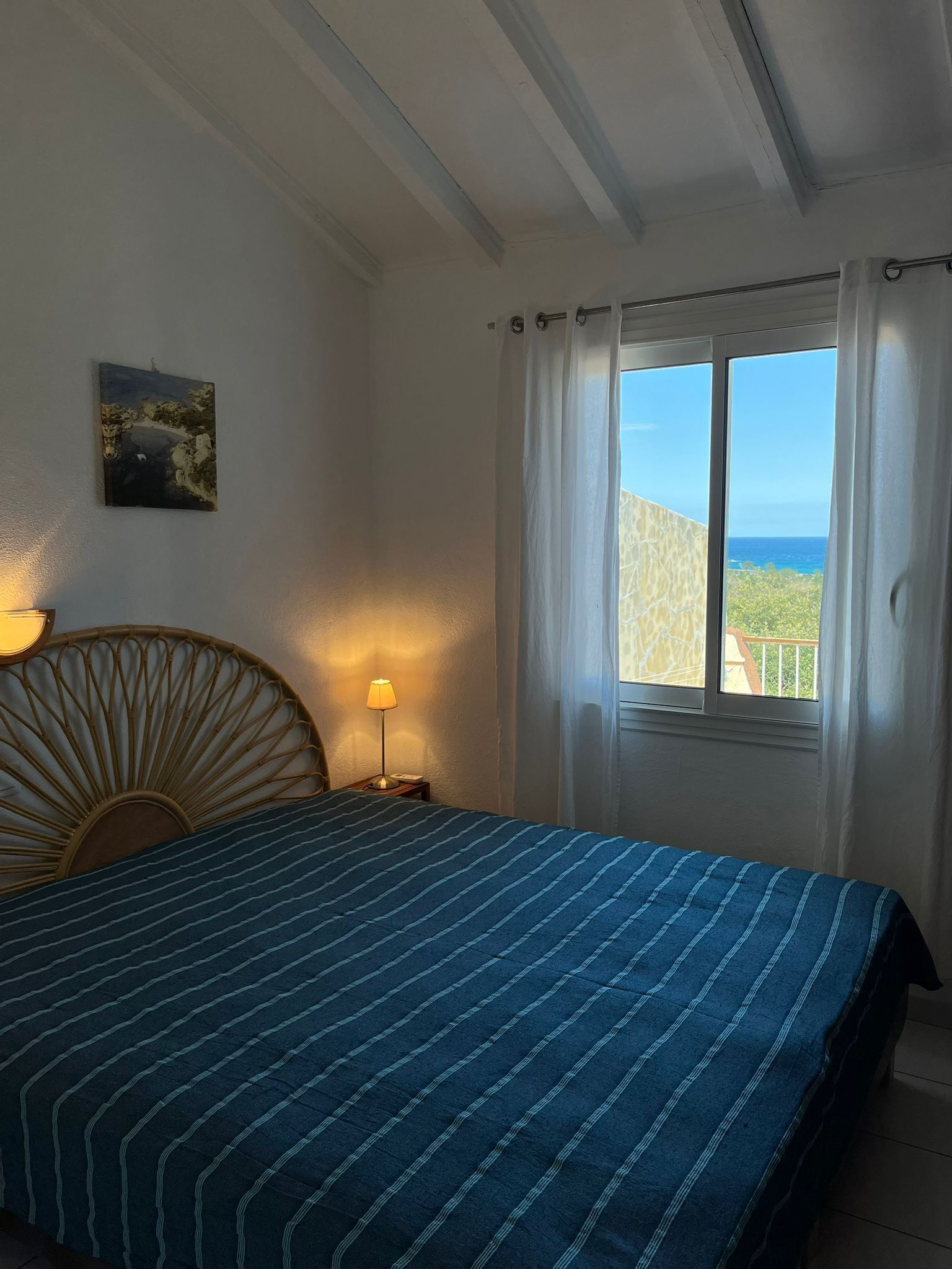 A bedroom with a bed, a nightstand with a lamp, a wall-mounted painting, and a window with a view of the sea.