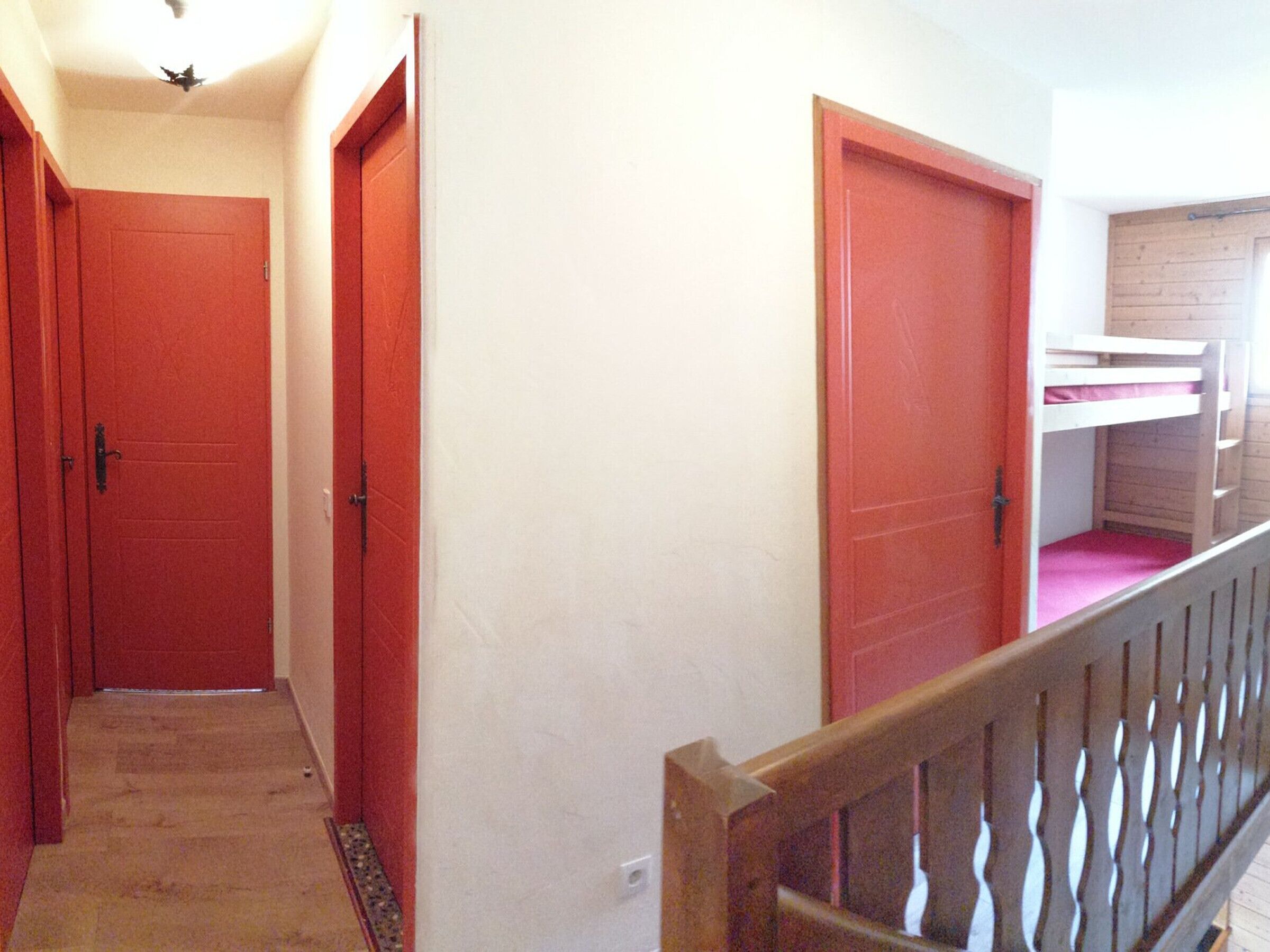 A hallway with red doors, a wooden railing, and a bunk bed in the background.