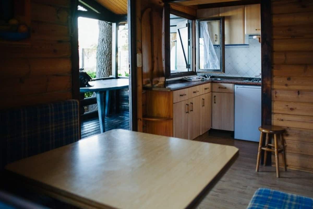 A wooden cabin interior with a dining table and chairs, a kitchen with wooden cabinets and a white refrigerator, and a window overlooking greenery.