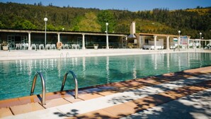 A swimming pool with metal railings, a covered seating area, and a forested hillside in the background.