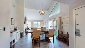 A spacious dining area with a wooden table and chairs, a television mounted above, and a large window providing natural light.