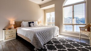 A bedroom with a bed, nightstand, lamp, and a view of the city through large windows.