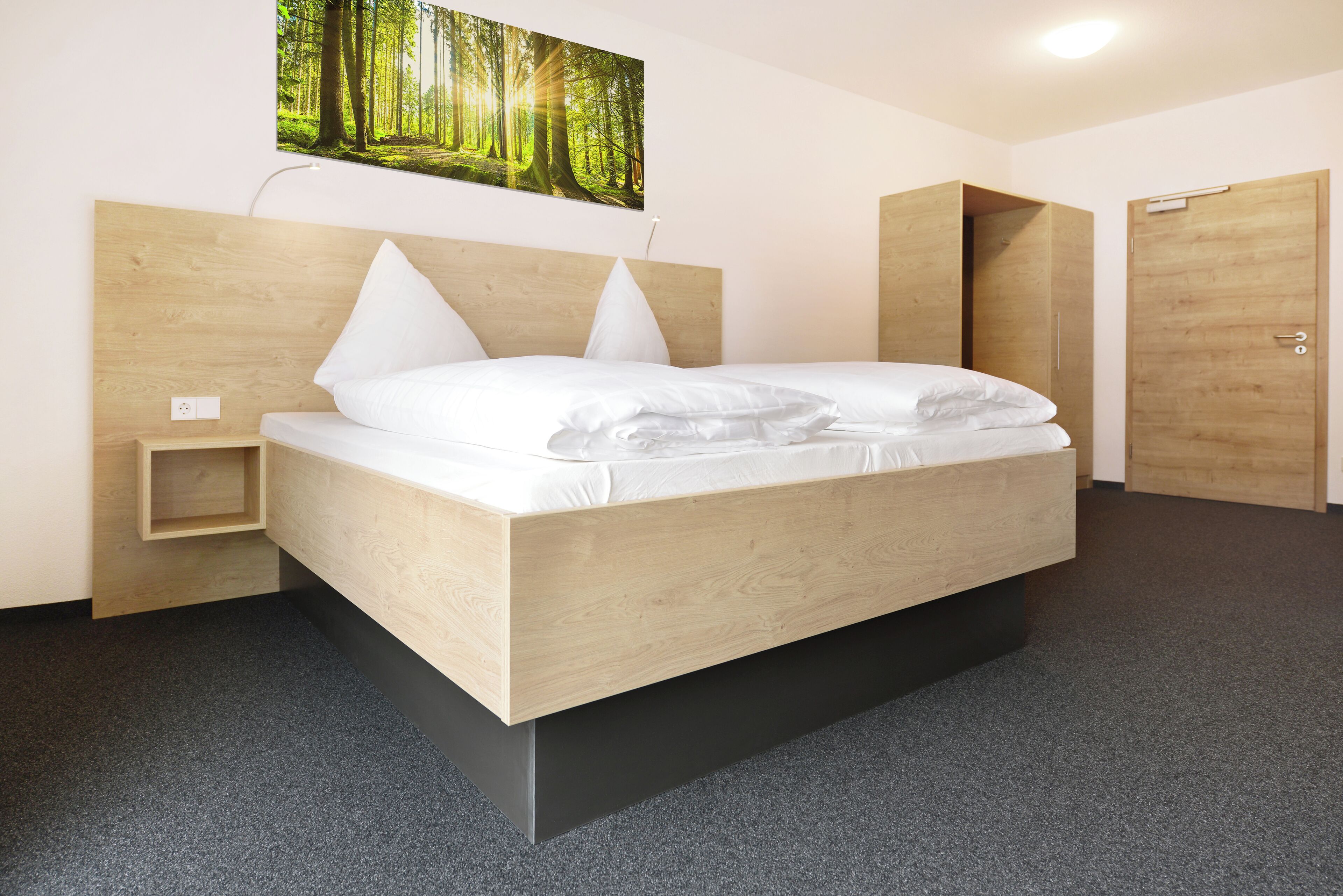 Un dormitorio moderno con cabecera de madera, cama con ropa blanca, una mesita de noche pequeña y un cuadro colgado en la pared con un paisaje forestal.