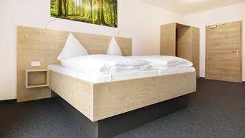 Un dormitorio moderno con cabecera de madera, cama con ropa blanca, una mesita de noche pequeña y un cuadro colgado en la pared con un paisaje forestal.