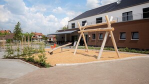 Ein Spielplatz mit Holzschaukeln und einer Wippe, umgeben von einer angelegten Grünfläche mit jungen Bäumen und einem Gebäude mit brauner Fassade und zahlreichen Fenstern.
