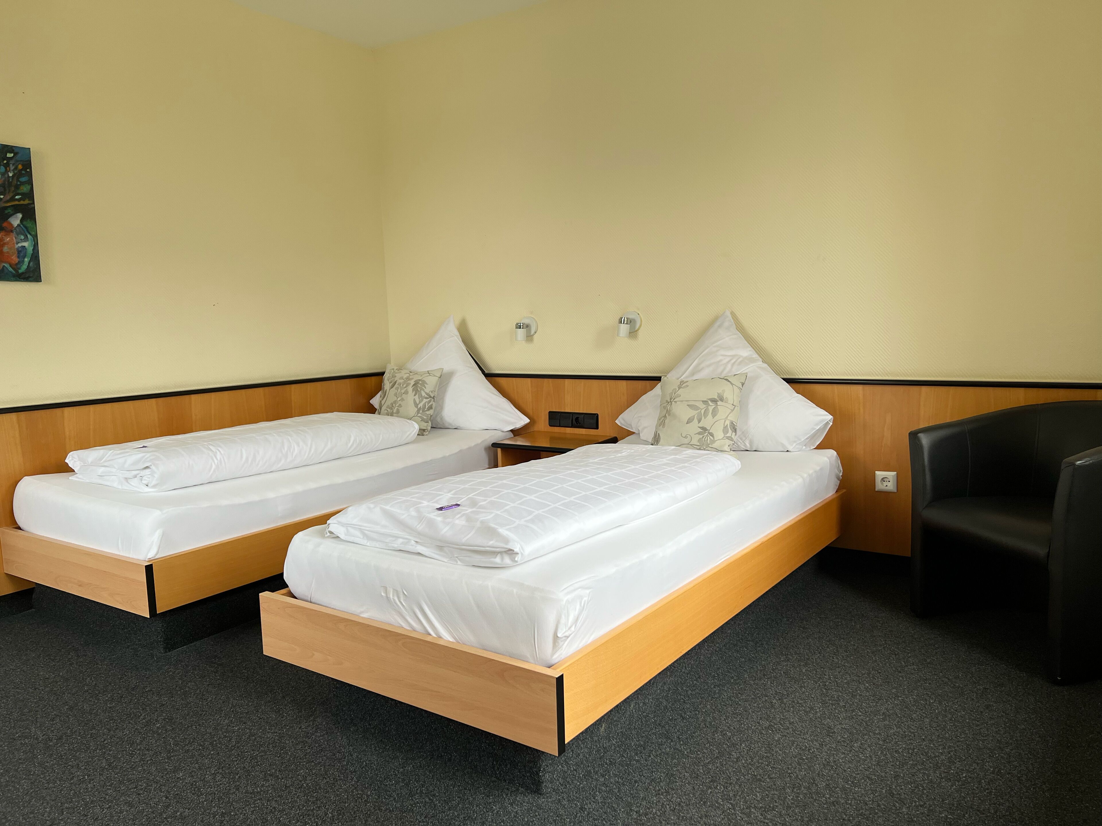 A hotel room with two single beds, a wooden headboard, a black armchair, and a small table.