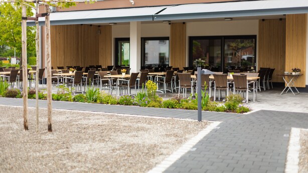 Ein Café mit Sitzbereich im Freien, Tische und Stühle, eine überdachte Lounge und ein Gebäude mit großen Fenstern und einem Balkon.